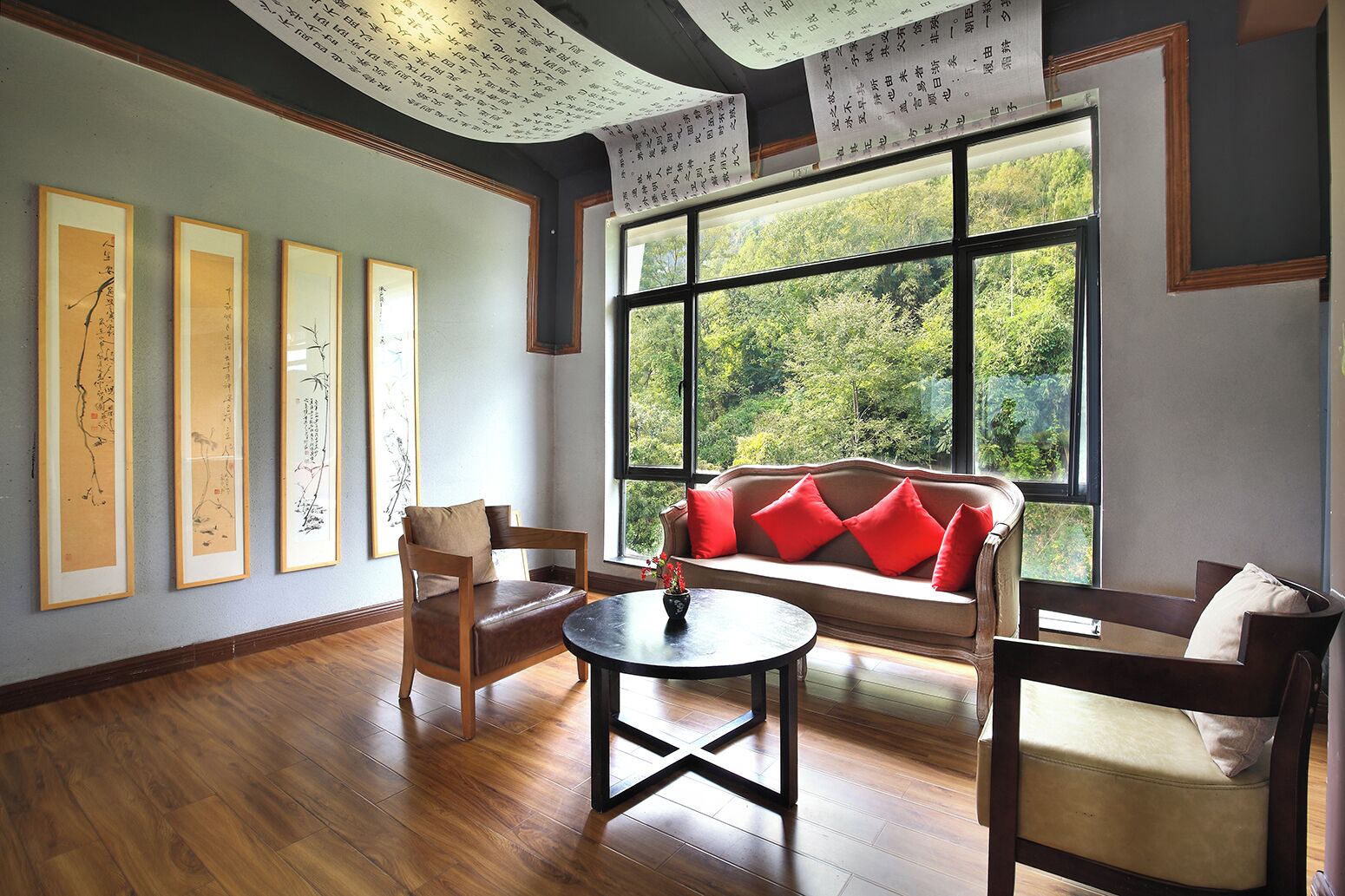 A room with a wooden floor, a sofa with red cushions, a wooden chair, a round table with a vase, and framed calligraphy on the wall.
