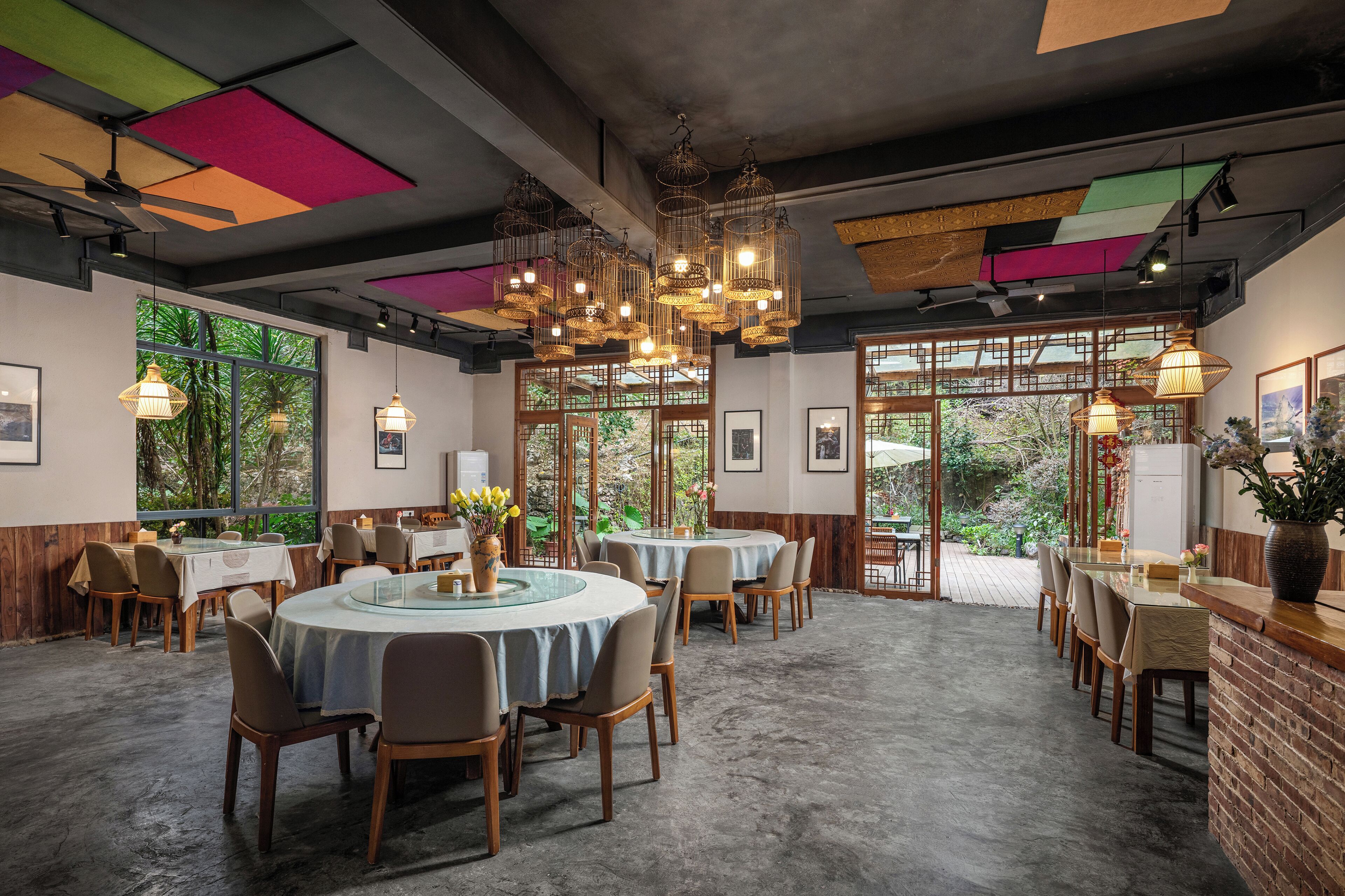Ein Restaurant mit runden Tischen, Stühlen, hängenden Lampen und großen Fenstern mit Blick auf Grünflächen.