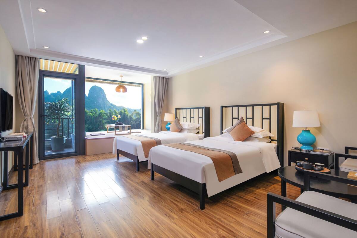 Habitación de hotel con dos camas, un escritorio, una silla, un televisor y un ventanal con vistas a las montañas.