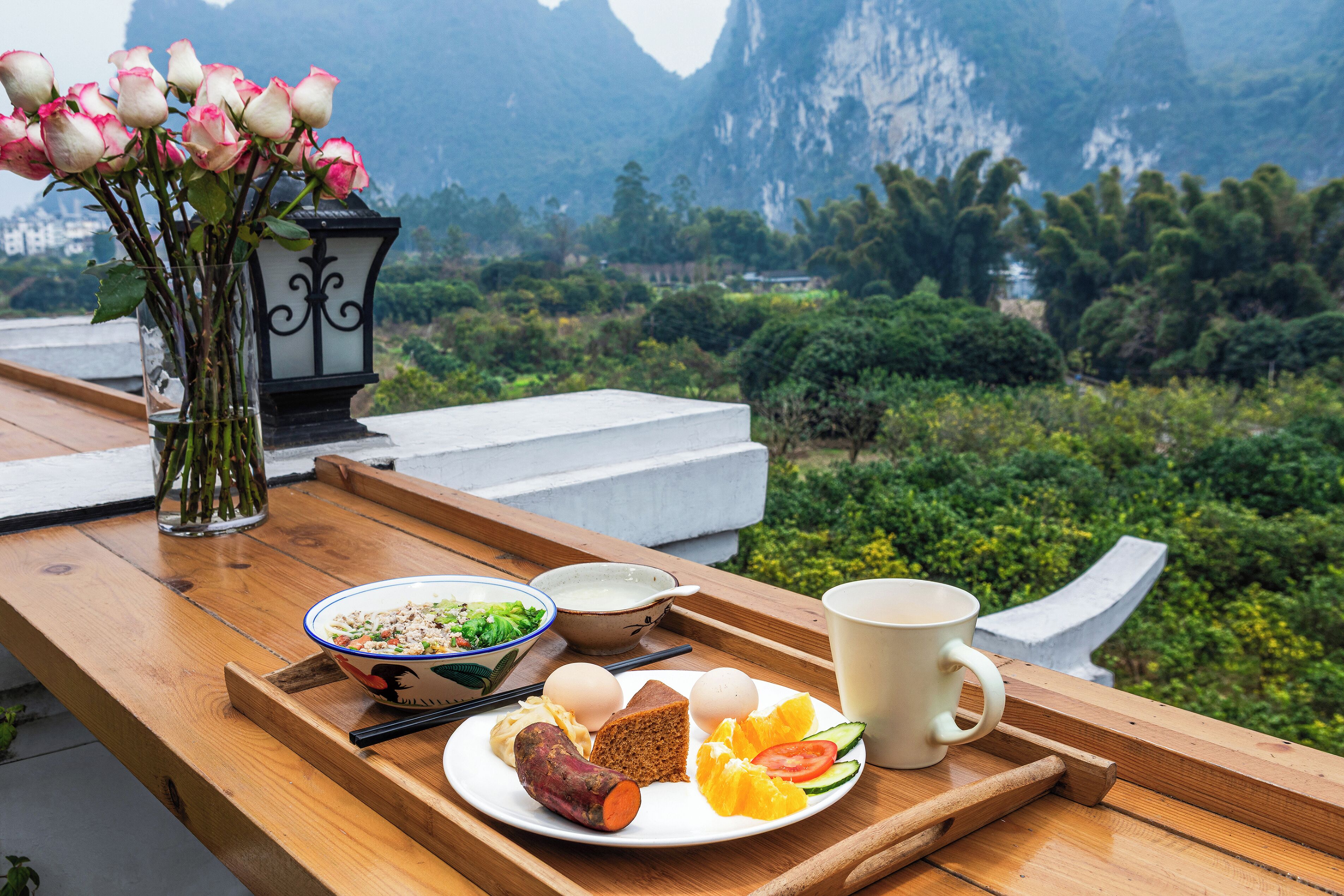 A wooden table with a meal, a vase of flowers, and a view of mountains.