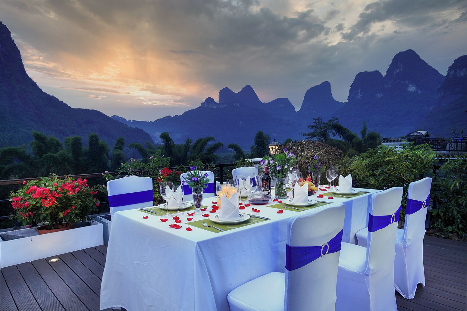 A table set for a meal with chairs, a view of mountains, and a balcony with potted plants.