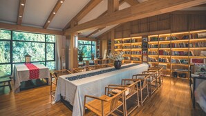 Une bibliothèque avec des étagères en bois, une longue table entourée de chaises et de grandes fenêtres.