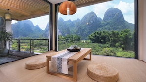 Une pièce avec une table en bois, deux poufs en fibres tressées, une grande fenêtre donnant sur les montagnes et un luminaire suspendu.