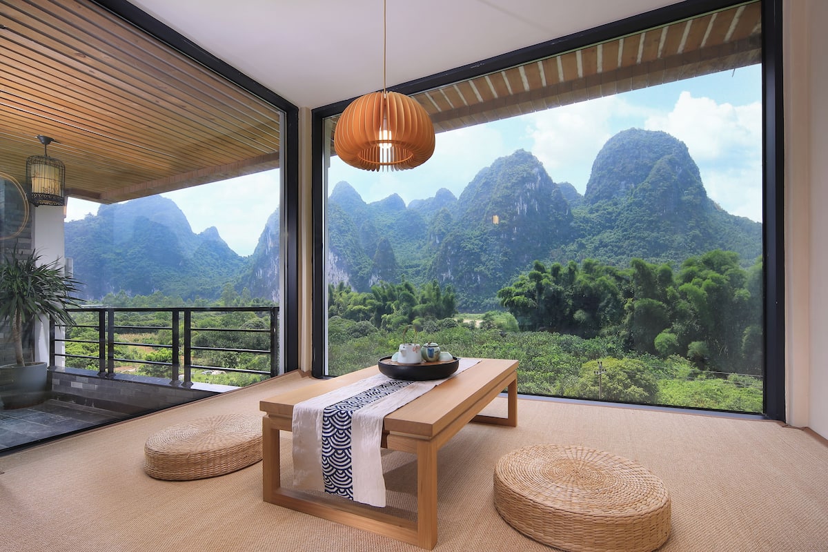 Una habitación con una mesa de madera, dos pufs tejidos, un candelabro colgante y un amplio ventanal con vistas a la montaña.