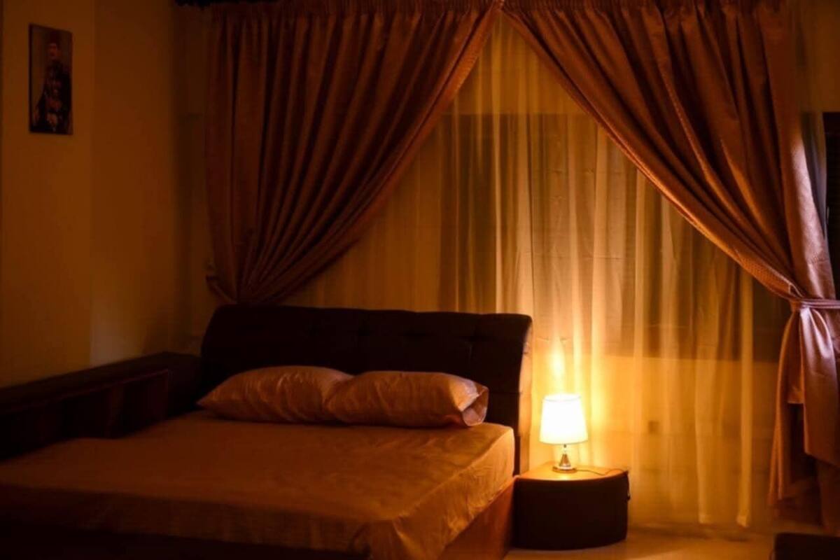 Un cuarto de hotel tenuemente iluminado con una cama, cortinas y una lámpara de noche.