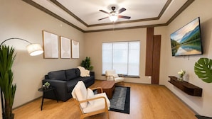 A living room with a dark sofa, a wooden chair, a coffee table, a ceiling fan, and a TV on the wall. - Downtown Tulsa Apartment Near Arts District  Pool (Tulsa)