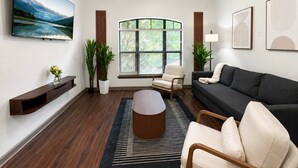 A living room with a TV, sofa, coffee table, and a window with blinds. - 2BR Modern Downtown Tulsa Stay Near BOK Center (Tulsa)