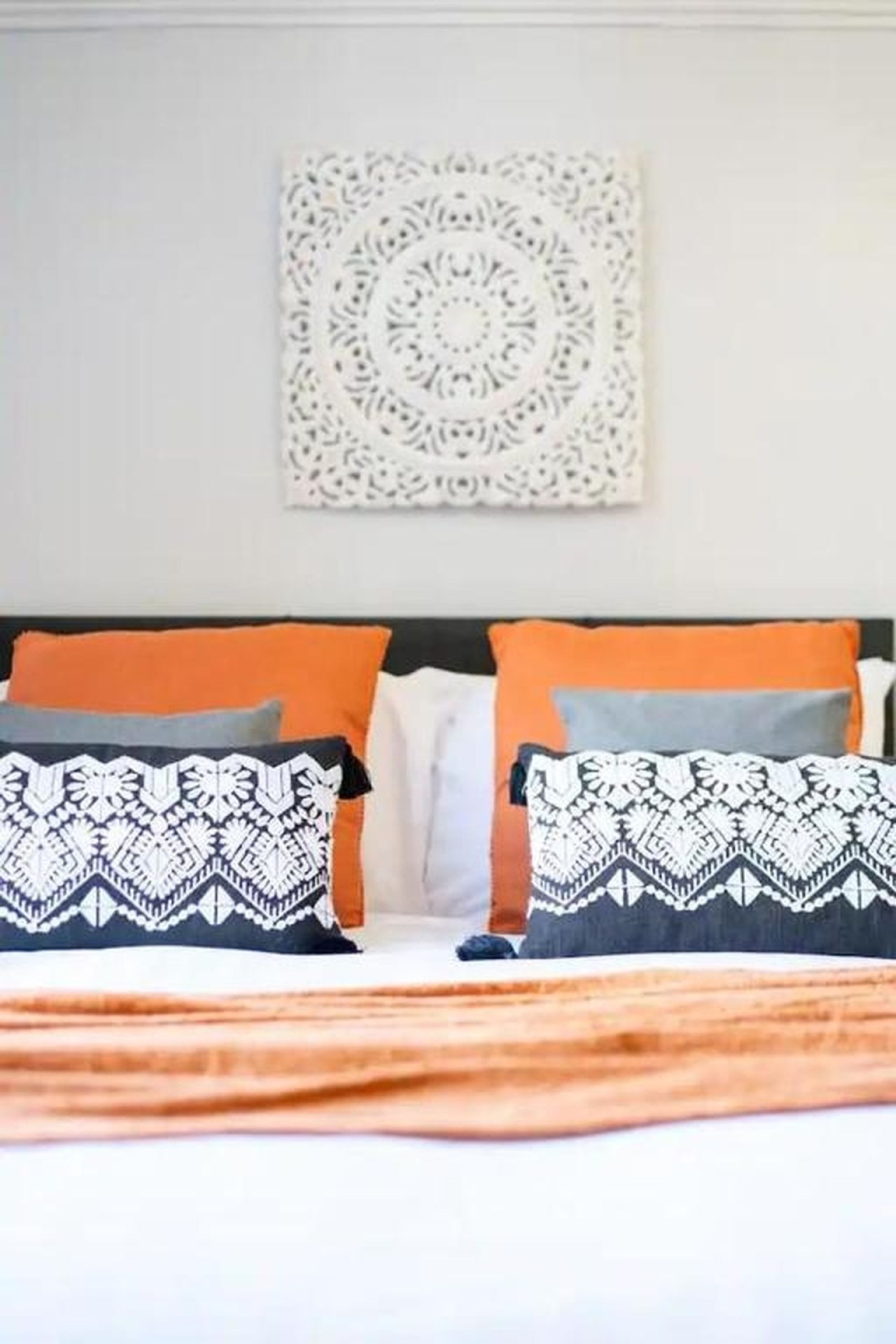A bed with patterned pillows, an orange throw blanket, and a wall-mounted decorative piece.