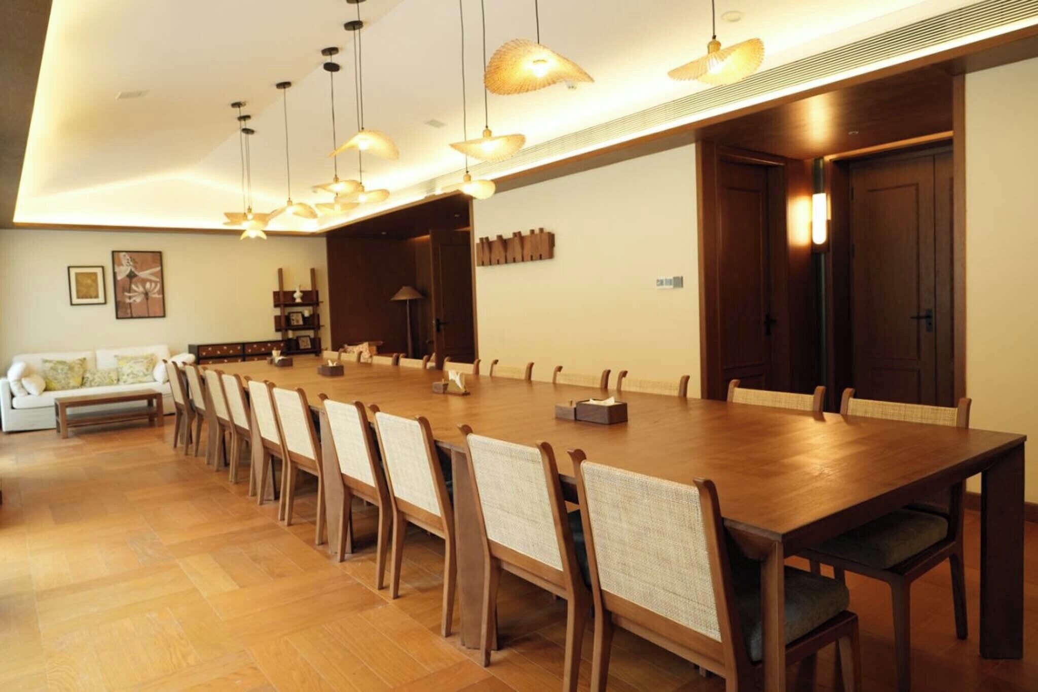 Una larga mesa de comedor con sillas, lámparas colgantes y un sofá en una habitación bien iluminada con piso de madera.