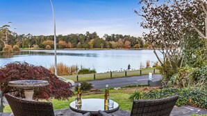 Un patio con mobiliario de mimbre, una mesa y una fuente para pájaros, con vistas a un lago con patos y un sendero peatonal.