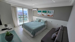 A modern bedroom with a bed, a sofa, a small table with a plant, and a view of buildings through the window. - Beach access | Sea view | Kitchen | WiFi | Comfort (Rio de Janeiro)