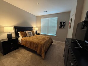Une chambre à coucher comprenant un lit, deux tables de chevet, une lampe, une fenêtre avec des stores et une commode.