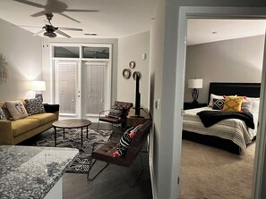 Ein modernes Wohnzimmer mit einer Couch, einem Couchtisch und einem Sessel. Ein Bett mit Kopfteil und Kissen ist ebenfalls vorhanden.