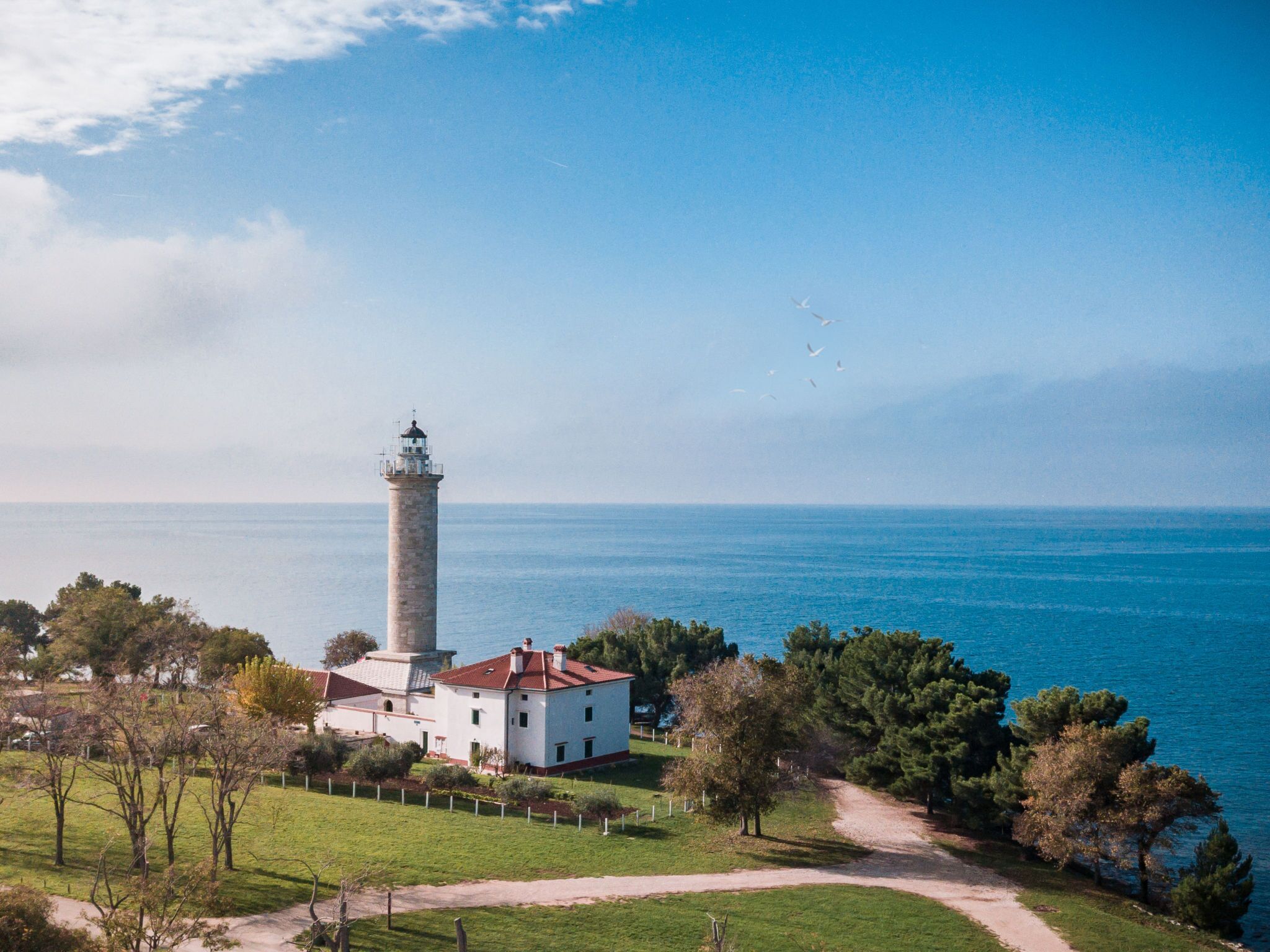 Ein Leuchtturm mit einem weißen Gebäude in der Nähe, umgeben von Bäumen und freiem Blick auf das Meer.