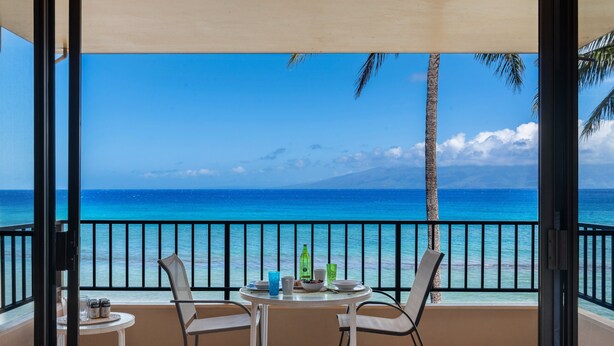 Een balkon met een tafel en stoelen, met uitzicht op een strand met helderblauw water en een berg in de verte.