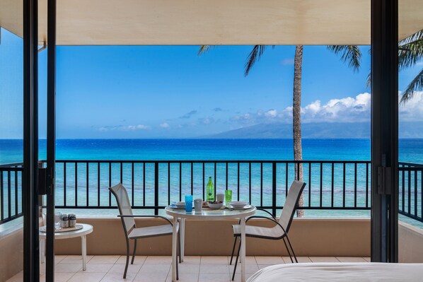 A balcony with a table and chairs, overlooking a beach with clear blue water and a mountain in the distance. - Ocean-view 2BR/2BA condo with pool, hot tub & beach access-Paki Maui 304 (Lahaina)