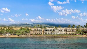 Hotel aan het strand met meerdere verdiepingen, palmbomen en uitzicht op de oceaan en bergen op de achtergrond.