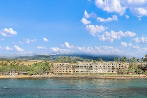 A beachfront hotel with multiple floors, palm trees, and a view of the ocean and mountains in the background. - Ocean-view 2BR/2BA condo with pool, hot tub & beach access-Paki Maui 304 (Lahaina)