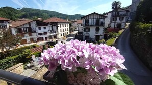 Ein Balkon mit Blumen, Blick auf ein Dorf mit traditioneller Architektur und eine Straße, die durch die Landschaft schlängelt.