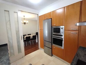 A compact kitchen with a refrigerator, microwave, and oven, wooden cabinets, a dining area with chairs, and a chandelier.