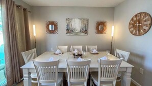 Un comedor con una mesa dispuesta para seis personas, rodeada de sillas blancas, con obras de arte y un reloj en la pared.
