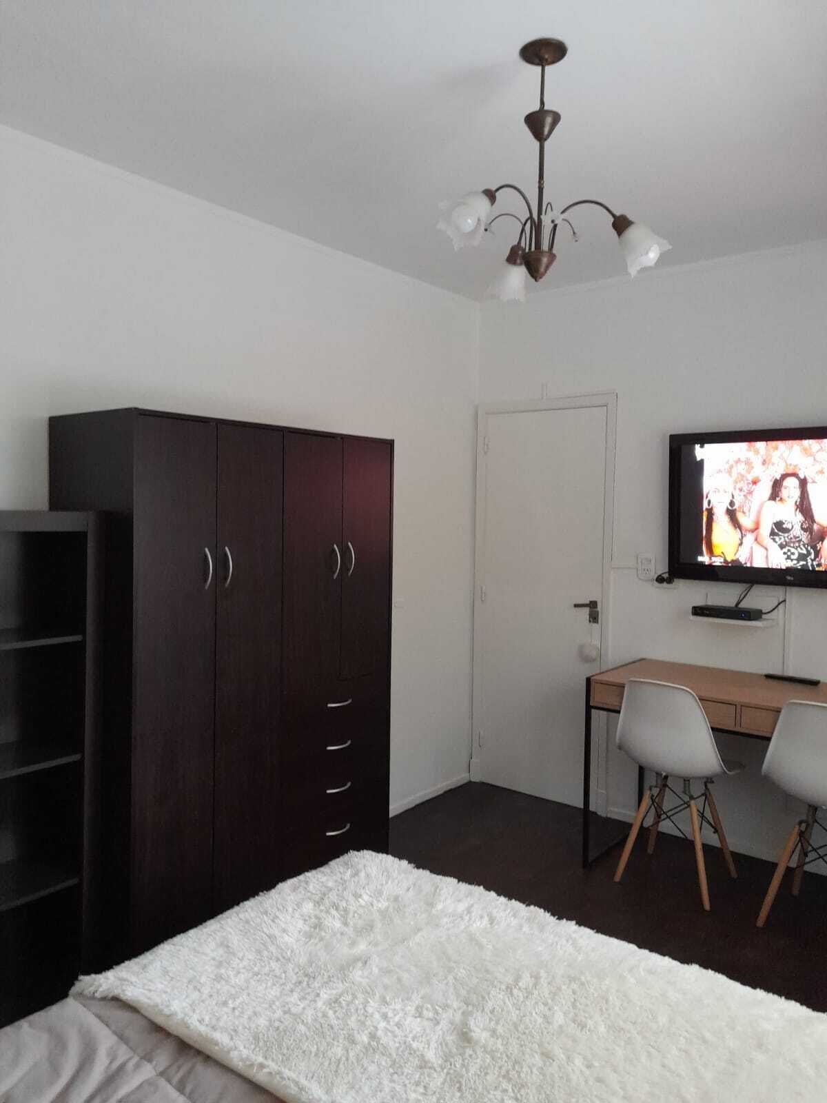 A bedroom with a bed, a wardrobe, a desk with two chairs, a television, and a ceiling light fixture.