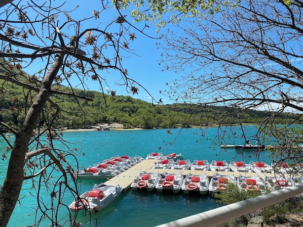 Ein Yachthafen mit vertäuten Booten, eine Holzsteganlage und im Hintergrund ein klarblauer See sowie grüne Hügel.