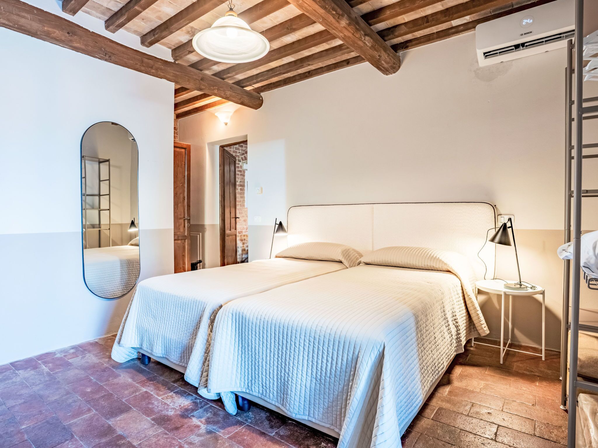 A double bed with a headboard, a bedside table with a lamp, a mirror, and a wooden ceiling with exposed beams.