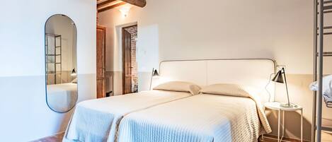 A double bed with a headboard, a bedside table with a lamp, a mirror, and a wooden ceiling with exposed beams.