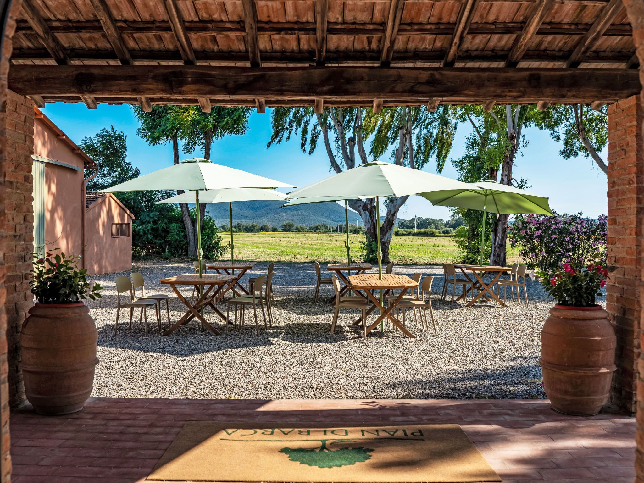 An outdoor patio with wooden tables and chairs, two umbrellas, and a view of a green landscape.