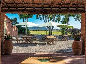 An outdoor patio with wooden tables and chairs, two umbrellas, and a view of a green landscape. - Apartment Madia by Interhome (Grosseto)
