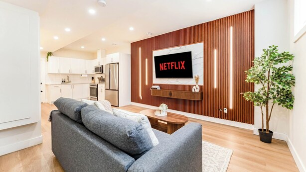 A modern living room with a grey sofa, wooden panel wall, a flat-screen TV, and a potted plant. - Entire Spacious Luxury Retreat Near DT | Sleeps 4 | Fast Wifi | Free Parking (Calgary)