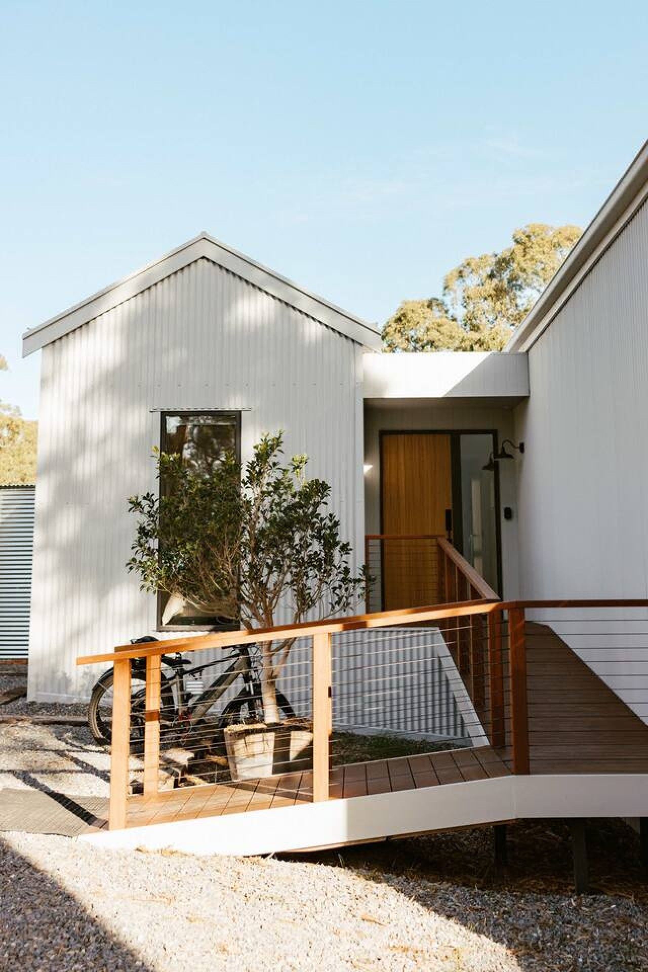 Une maison moderne de plain-pied, avec un escalier en bois menant à la porte d’entrée, un petit porche et un vélo garé à l’extérieur.