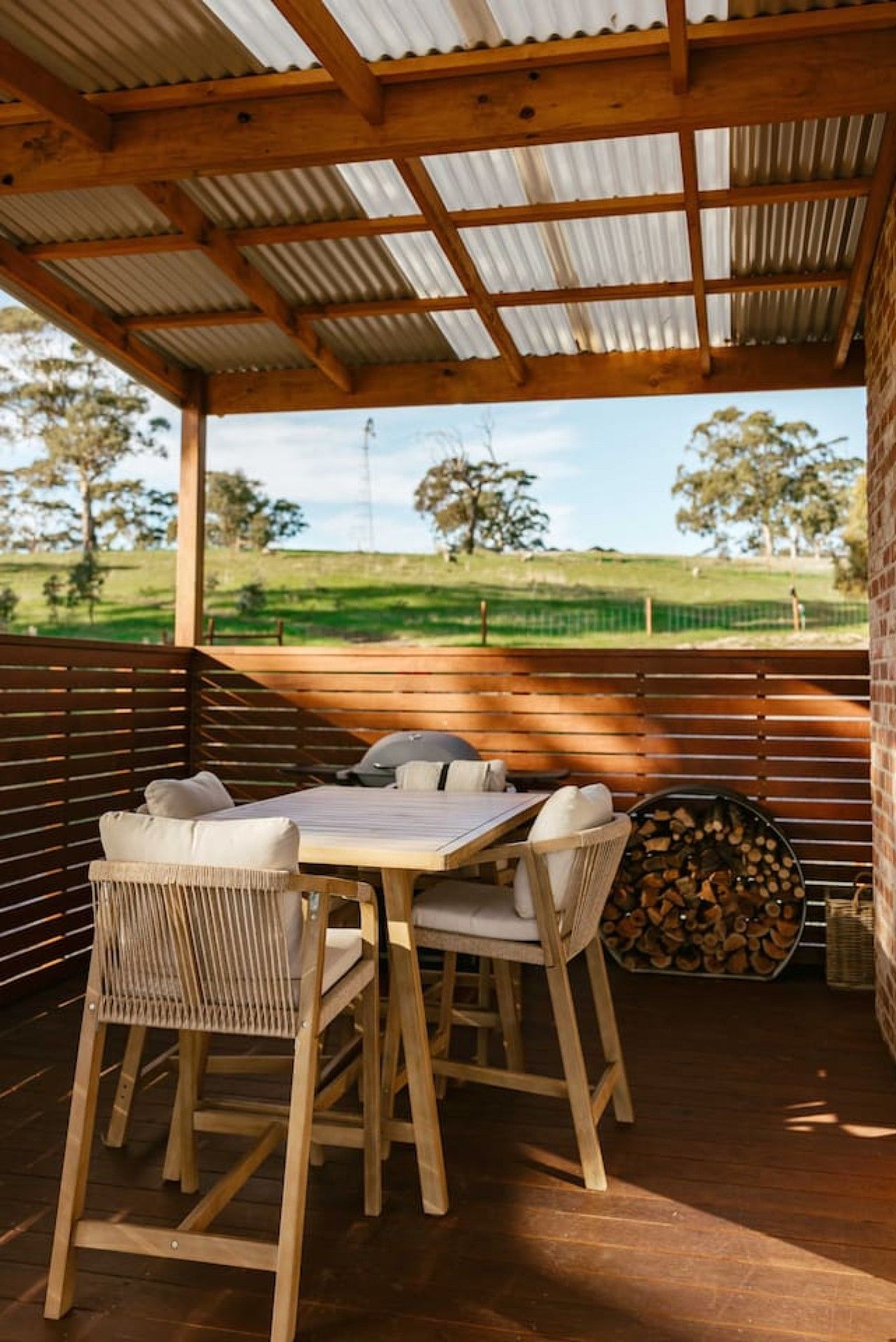 An outdoor patio with a wooden table and chairs, a grill, and a stack of firewood.