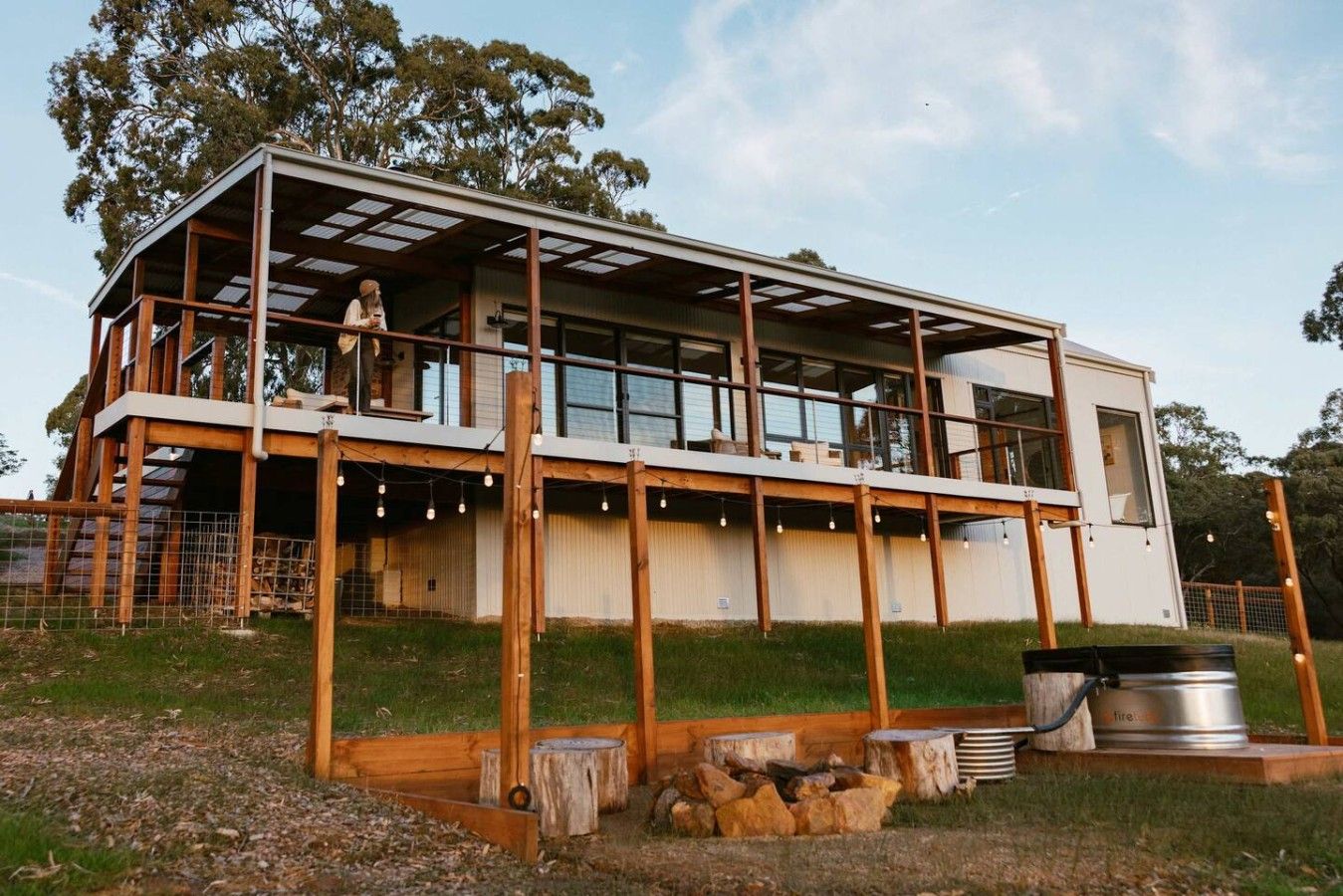 A modern two-story house with a large balcony, wooden deck, and a fire pit area.