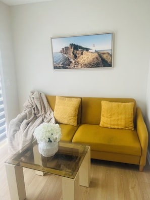 Ein Wohnzimmer mit einem gelben Sofa, einem Glastisch, einer weißen Vase mit Blumen, einer beigen Tagesdecke und einem Gemälde von einer Küstenlandschaft an der Wand.