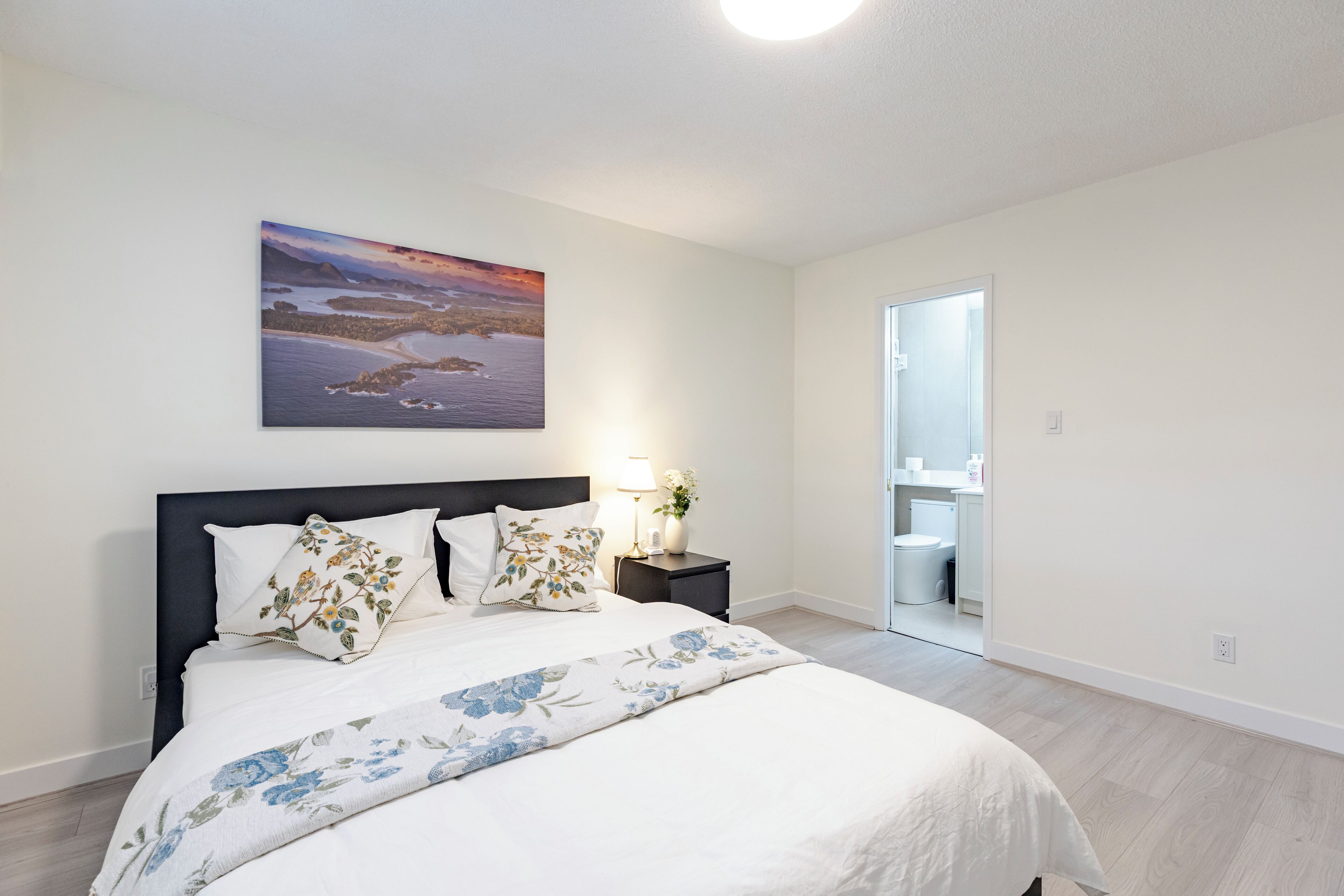 A bedroom with a bed, a nightstand, a lamp, a painting, and a bathroom visible through an open door.