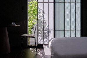 A room with a bed, a chair, a small table, and a view through a window with a bamboo plant outside.