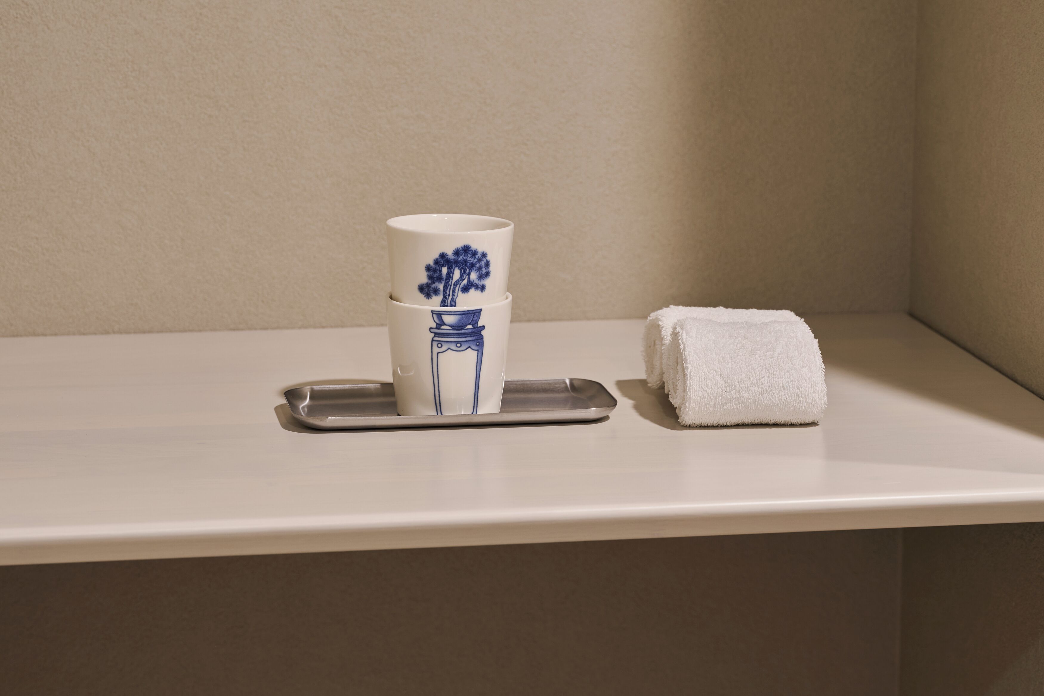 Een stapel witte bekers met blauwe patronen op een dienblad, een witte handdoek en een beige muur.