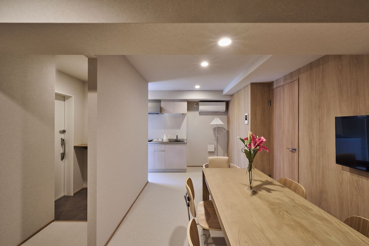 A modern kitchen with white cabinets, a dining area with a wooden table and chairs, and a TV mounted on the wall.