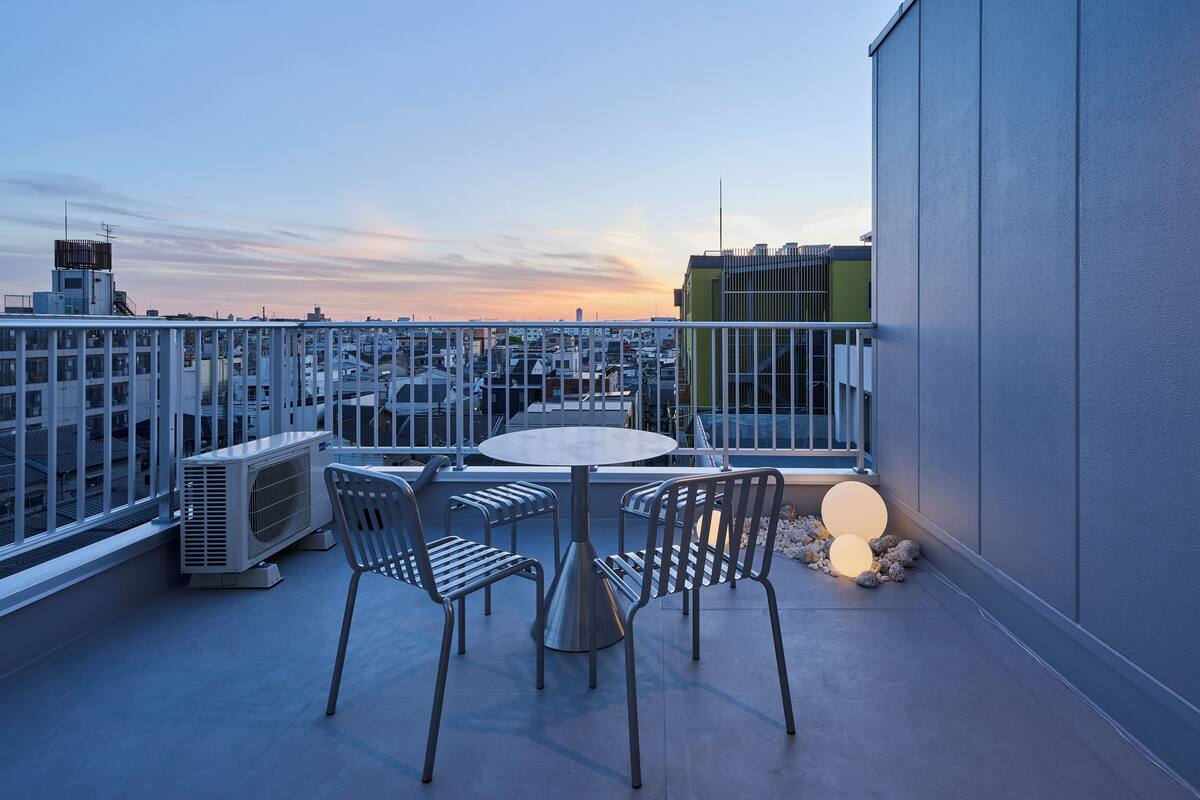 A rooftop terrace with a round table and chairs, air conditioning units, and a view of the cityscape.