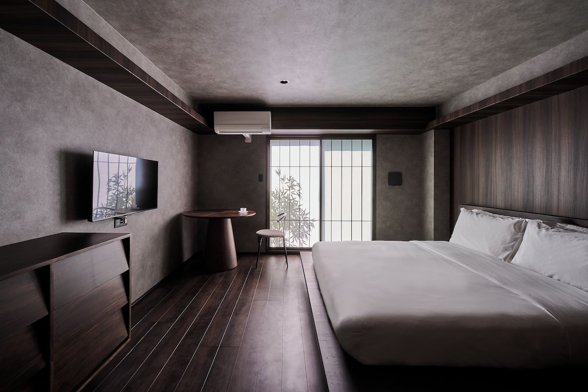A modern hotel room with a large bed, a TV mounted on the wall, a small round table, and a window with a view of trees.