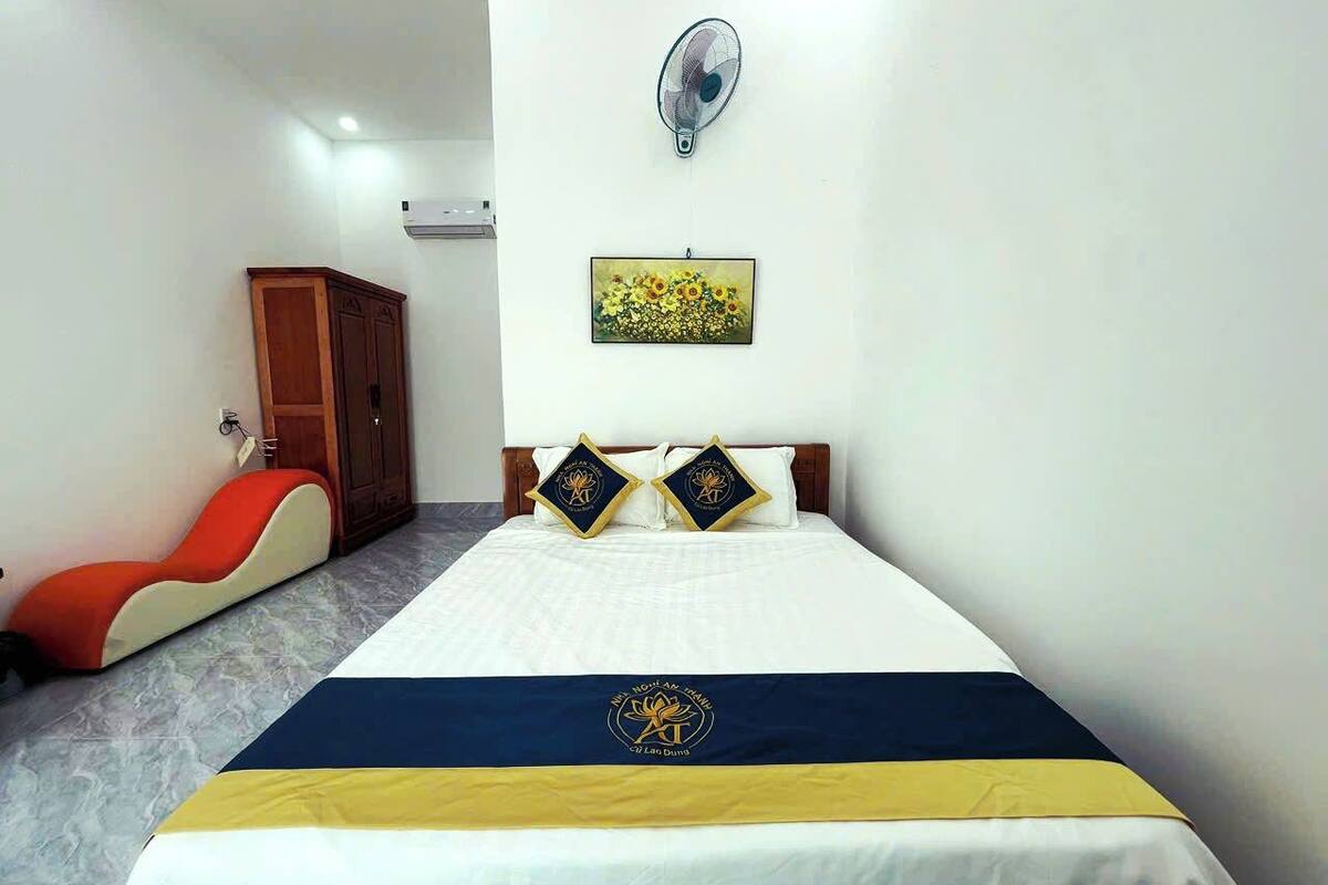 Una habitación de hotel con cama, ventilador de pared, un cuadro y una silla roja.