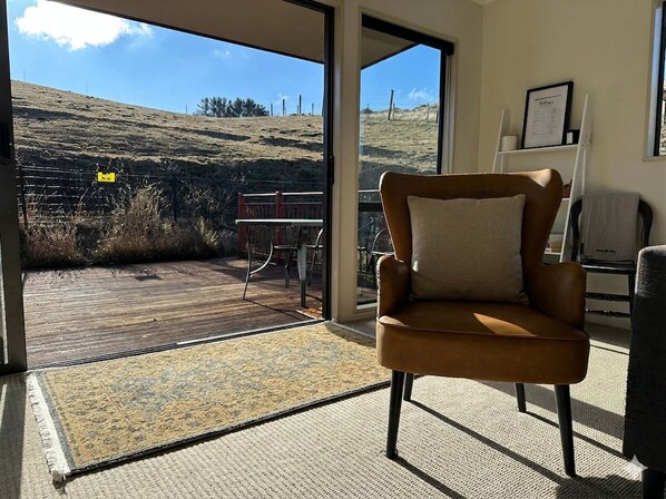 A room with a brown leather chair, a large window with a view of a grassy hill, and a door leading outside. - Clydesvale Hideaway (Springvale)