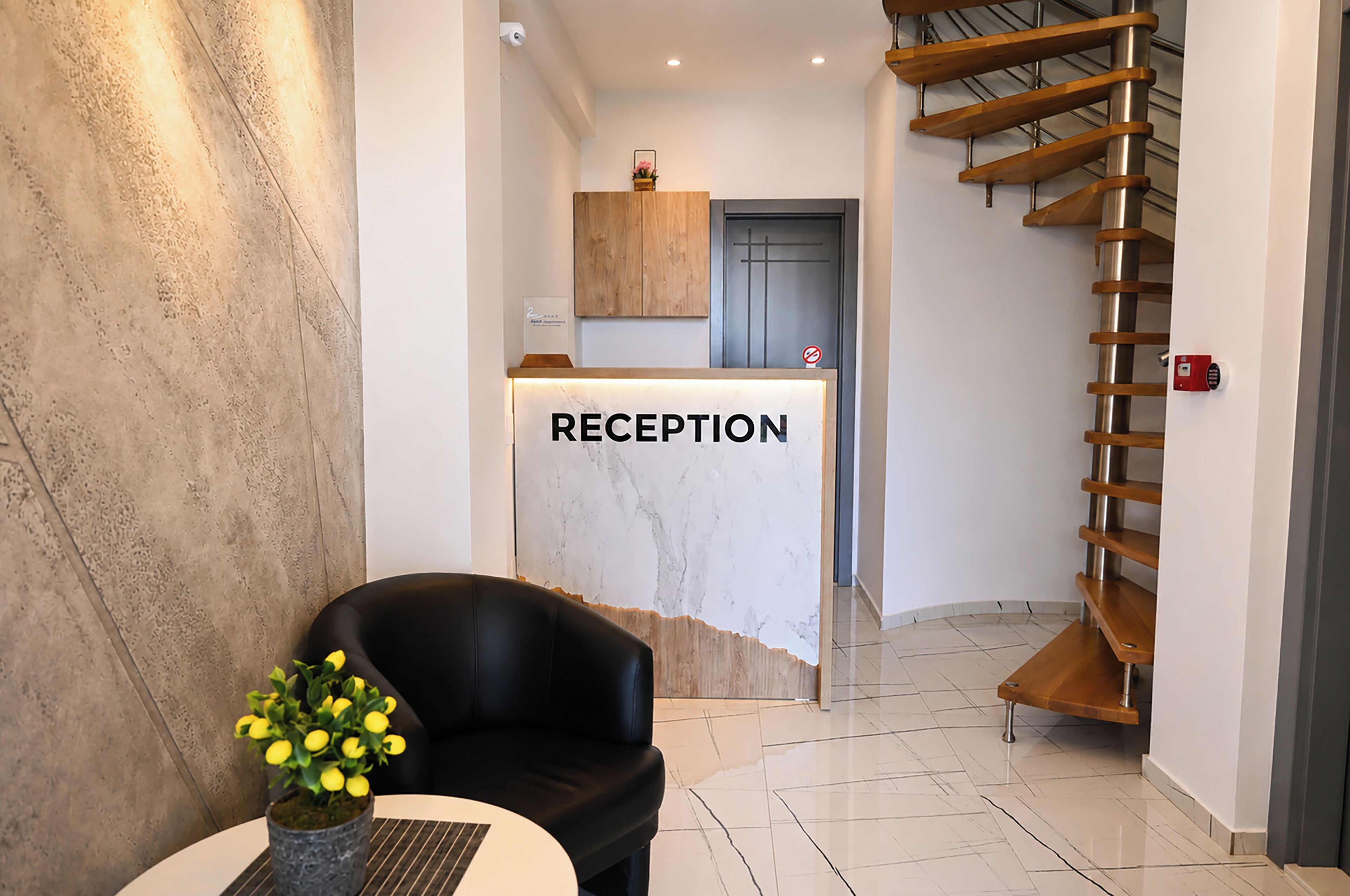 Een receptiebalie met de tekst 'RECEPTION', in een modern interieur met een marmeren wand, een zwarte lederen fauteuil, een houten trap en een tafeltje met een vaas gele bloemen.