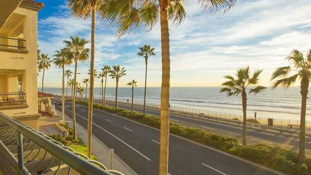 A balcony with a view of a coastal road lined with palm trees and a beach. - Unwind In Premium Beach-front Resort with Panoramic Ocean Views. 1Bd w/ Balcony (Carlsbad)