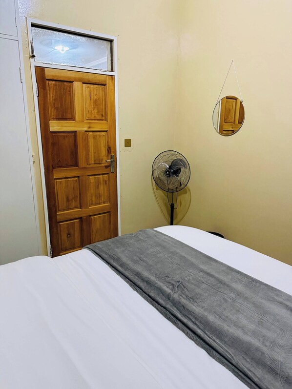 A room with a wooden door, a bed with a white and gray blanket, a standing fan, and a mirror on the wall.
