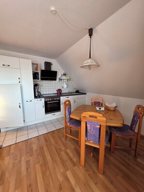 A compact kitchen with white cabinets, a wooden dining table with chairs, a hanging light fixture, and a tiled backsplash. - Vacation apartment Auszeit-Anker. A place for people and animals to relax. (Werdum)
