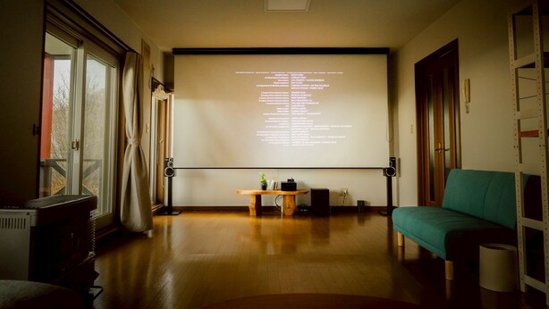 Una habitació amb pantalla de projector que mostra text, un sòl de fusta, un sofà i una taula amb una planta.