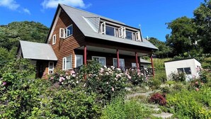 Una casa de dos pisos amb teulada inclinada, un porxo i un jardí amb diverses plantes i flors.